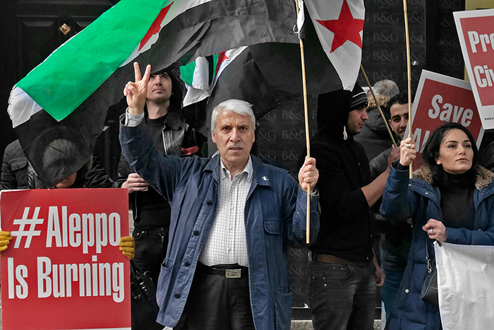 Un vieil homme tient un drapeau syrien d'une maine et fait le V de la victoire de l'autre main. Une pancarte à côté de lui : "Aleppo is burning"