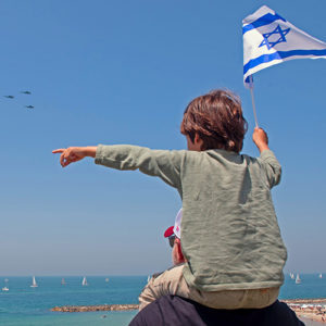 Enfant sur les épaules de son père tient u ndrapeau israélien dans sa main droite et avec la gauche indique les avions acrobatiques de l'armée israélienne