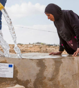 Femme palestinienne qui regarde l'eau couler dans un réservoir avec un panneau qui dit qu'il a été construit grâce à un financement de l'Union européenne