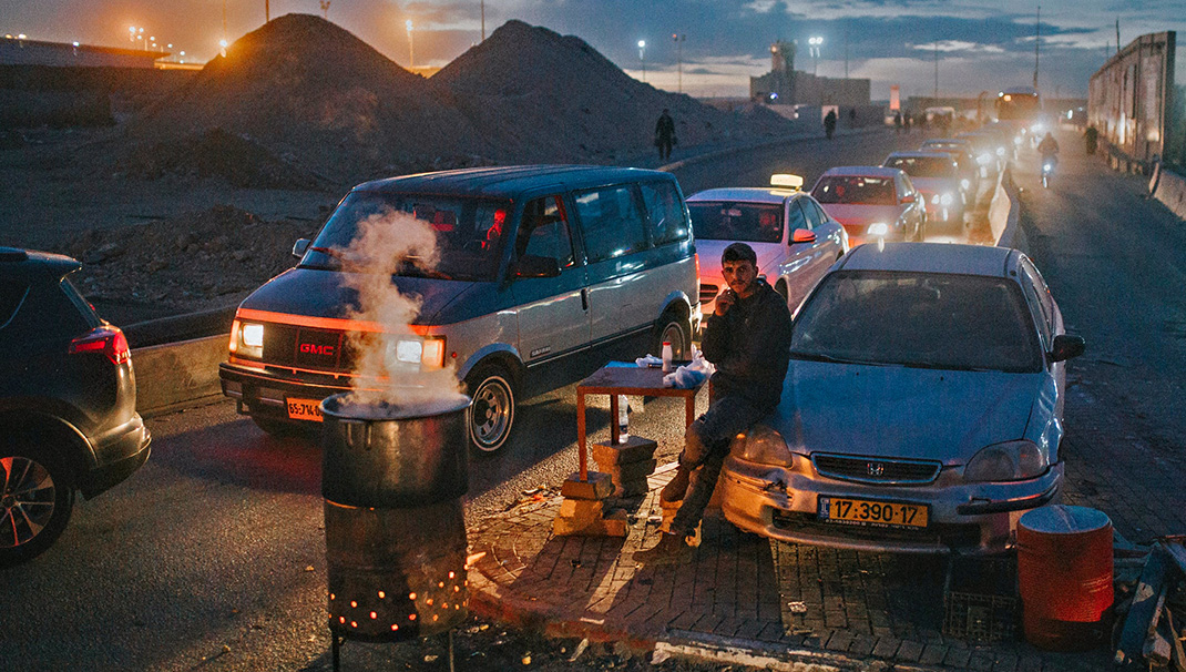 jeune palestinien à un check-point israélien se chauffe la nuit devant un feu; Une longue file de voiture derrière lui en attente de passer les contrôles