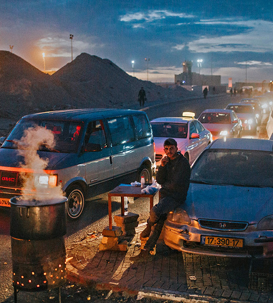 jeune palestinien à un check-point israélien se chauffe la nuit devant un feu; Une longue file de voiture derrière lui en attente de passer les contrôles