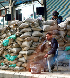 deux soldats syriens montent la garde. le premier se trouve derrière la barricade faite de sacs de sable, alors que le premier, assis sur une chaise, regarde son téléphone
