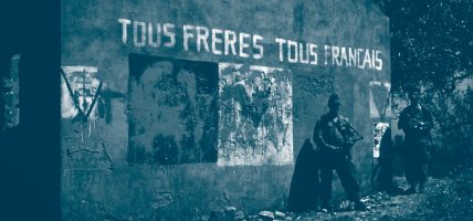 Deux algériens armés en uniforme militaire posent devant un graffiti représentant le drapeau français tâché avec un text qui dit "Tous frères, tous Français"